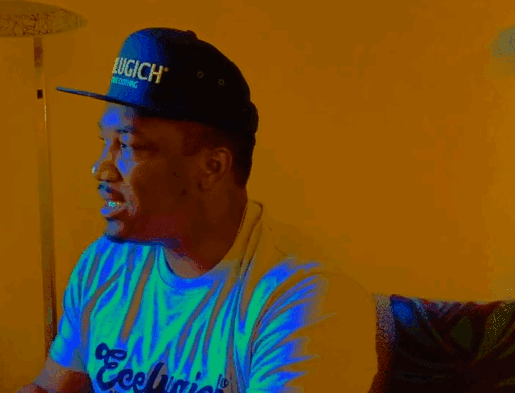 Man in a cap and graphic tee looking to the side against an orange-yellow backdrop.